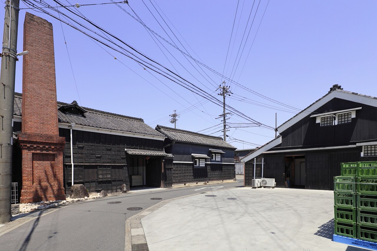 名古屋市緑区大高町の萬乗醸造。晴れた空の下、黒い板壁と瓦屋根の建物が道沿いに並び、左手にれんが造りの煙突が立つ。右手の建物の前には緑色のケースが積まれている