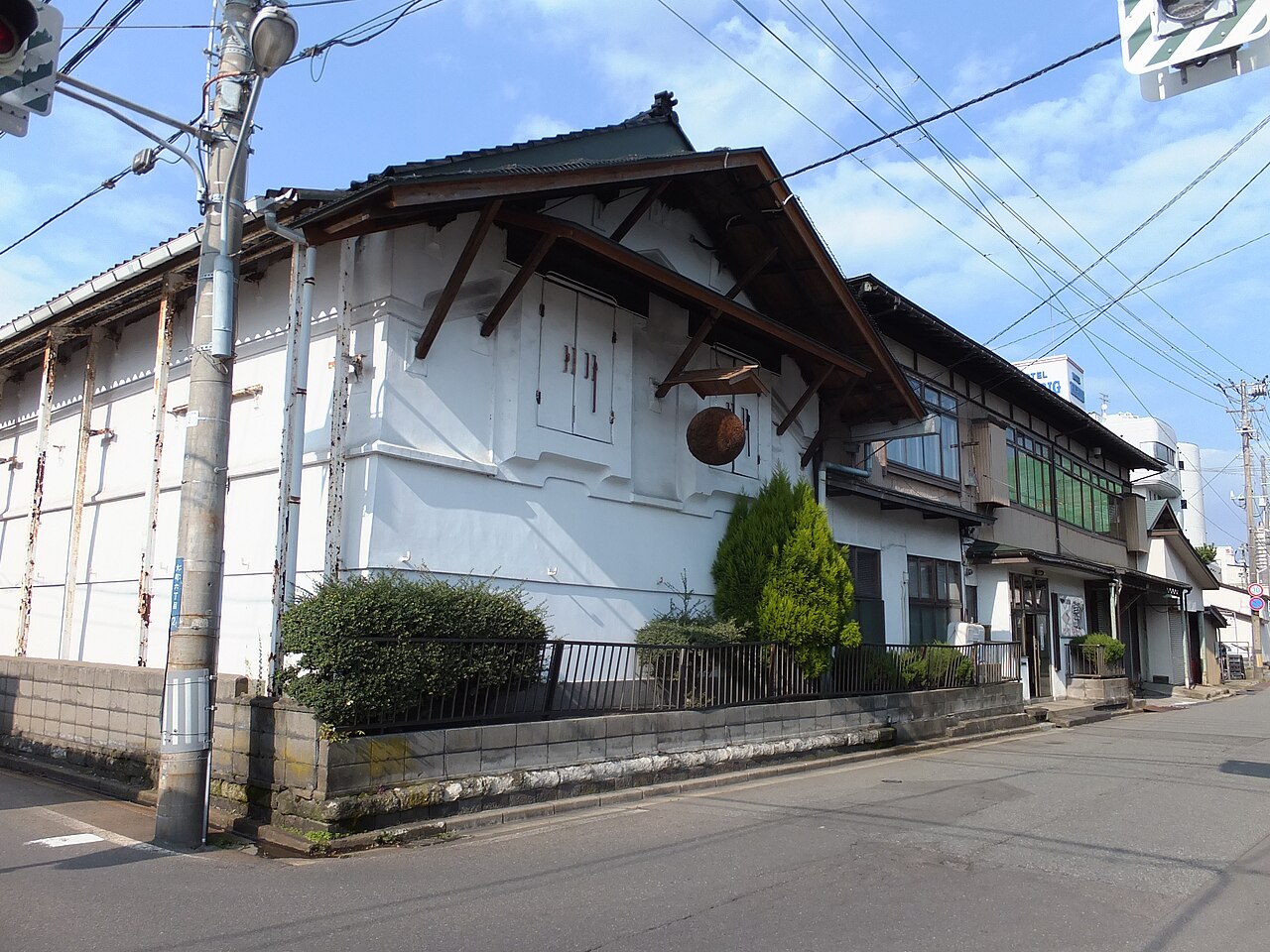 晴れた日の新政酒造の建物。白い壁の蔵の軒下に茶色い杉玉が下がり、右手に木造の建物が続く。手前は道路と低い塀で、電線と電柱が写る