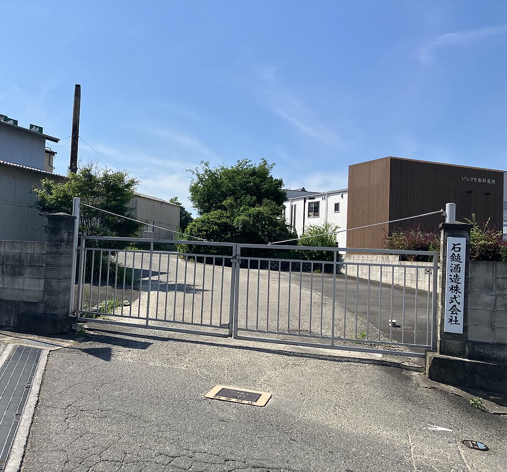 石鎚酒造の閉じた鉄の門と、門柱の「石鎚酒造株式会社」の表札。奥に蔵の建物と煙突、木々が見える