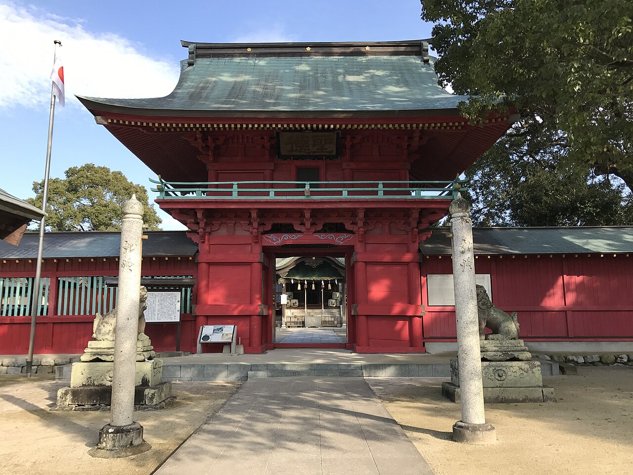 晴れた日の北野天満宮の楼門。緑青色の屋根をのせた朱塗りの二層の門が正面に立ち、手前に石畳の参道、その両脇に狛犬と石柱がある。門の奥に拝殿が見える