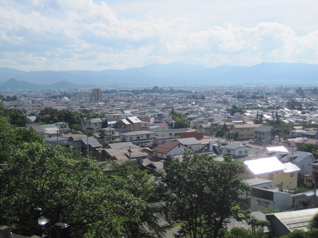 夏の飯盛山から見下ろした会津若松の市街。手前に木々、その先に住宅の屋根が広がり、遠くに盆地を囲む山並みがかすんで見える