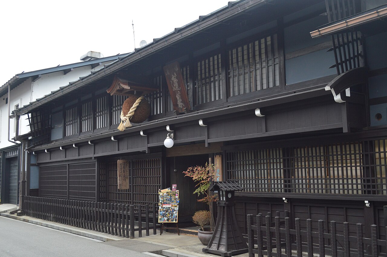 飛騨古川の壱之町に建つ渡辺酒造店の主屋。黒っぽい木の格子が続く2階建ての町家で、軒下に小さな屋根をかけた大きな杉玉が下がる