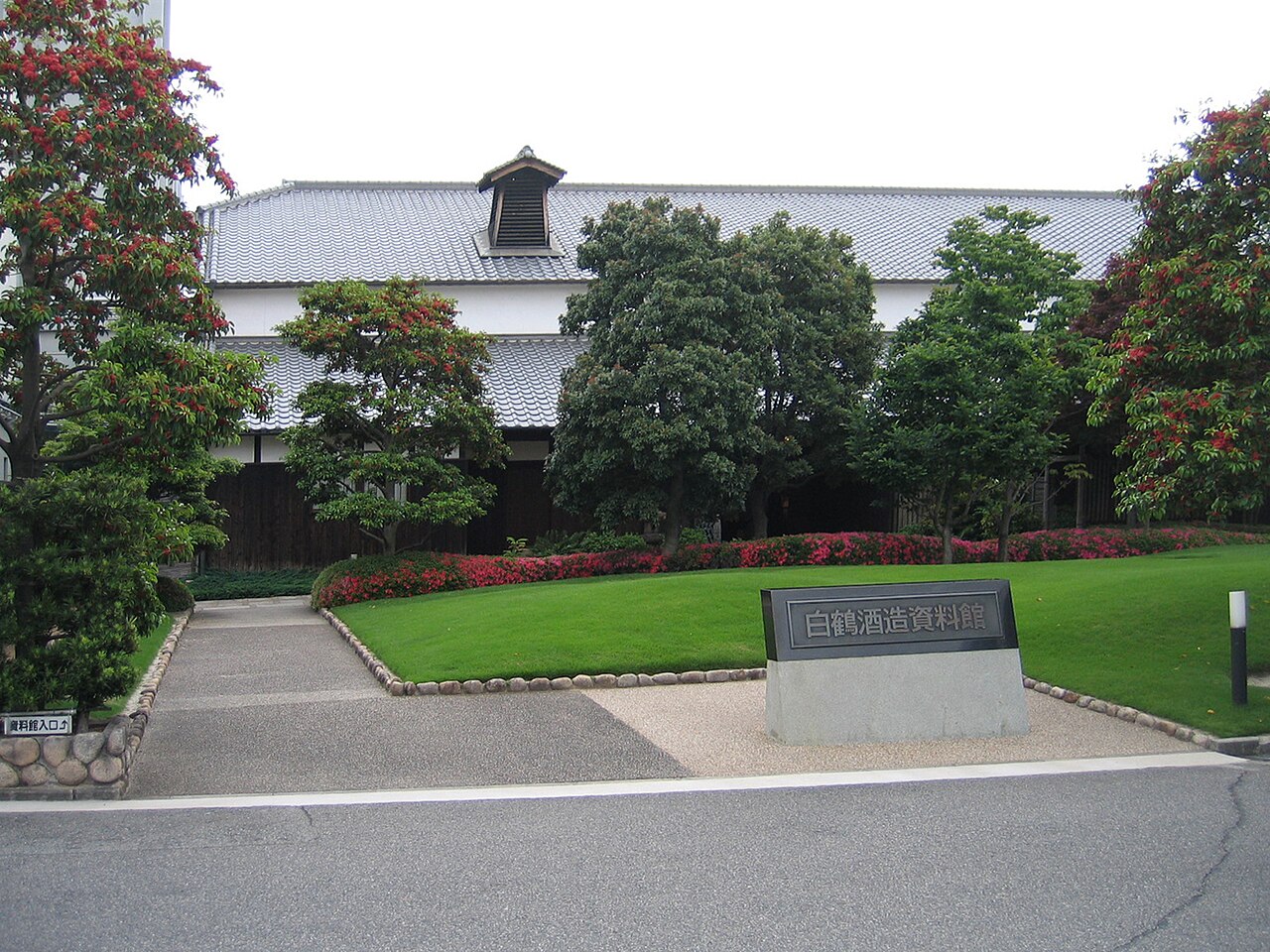 神戸市東灘区の白鶴酒造資料館。瓦屋根に煙出しの小屋根が載った白壁の木造の蔵が木々の奥に建ち、手前の芝生の脇に「白鶴酒造資料館」と刻んだ石の銘板が置かれている