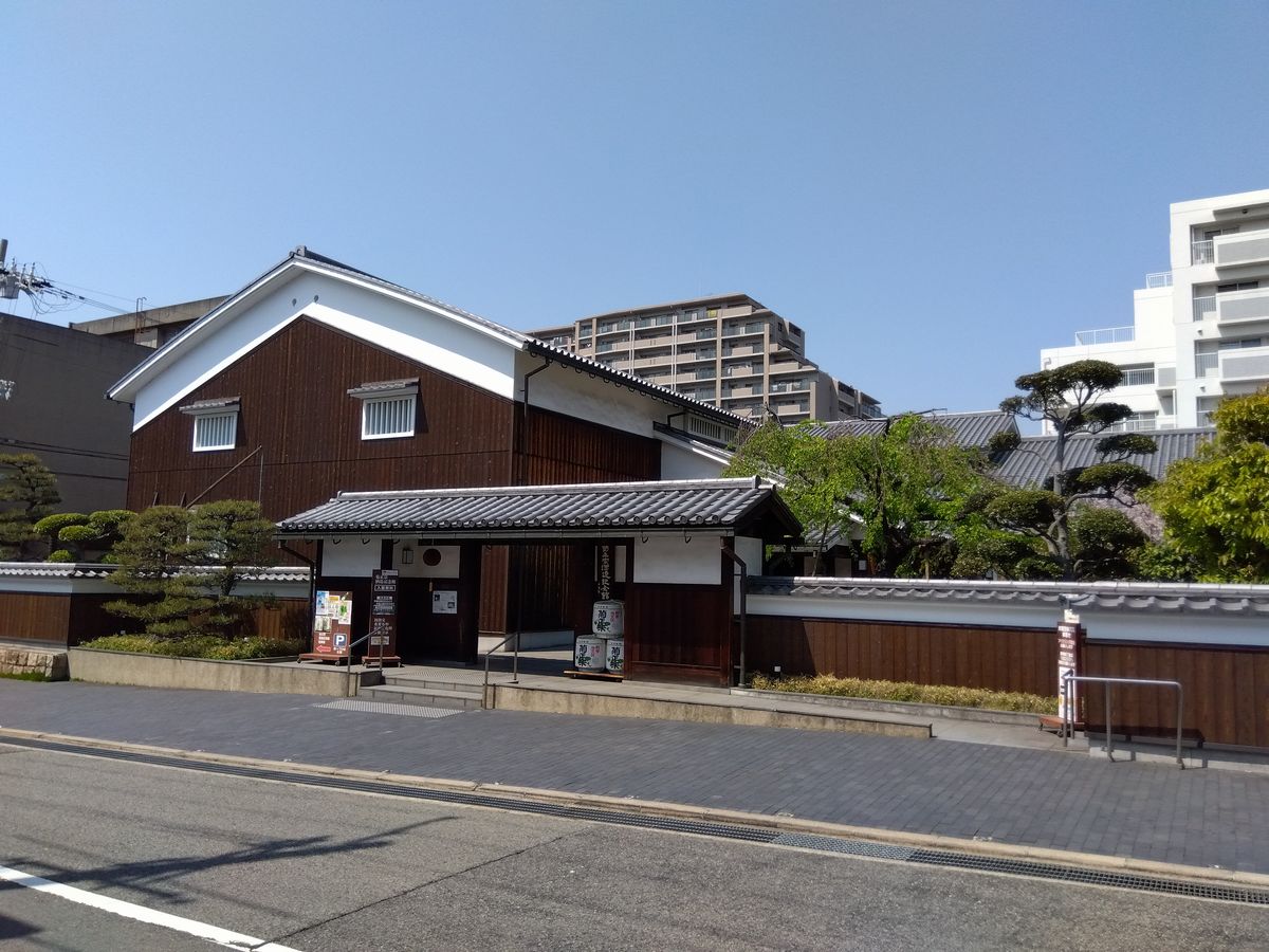 神戸市東灘区魚崎西町の菊正宗酒造記念館を道路の向かいから見た外観。白壁と焼杉板の蔵造りの建物に瓦葺きの門があり、門の脇に菰樽が積まれている。背後にはマンションが建つ