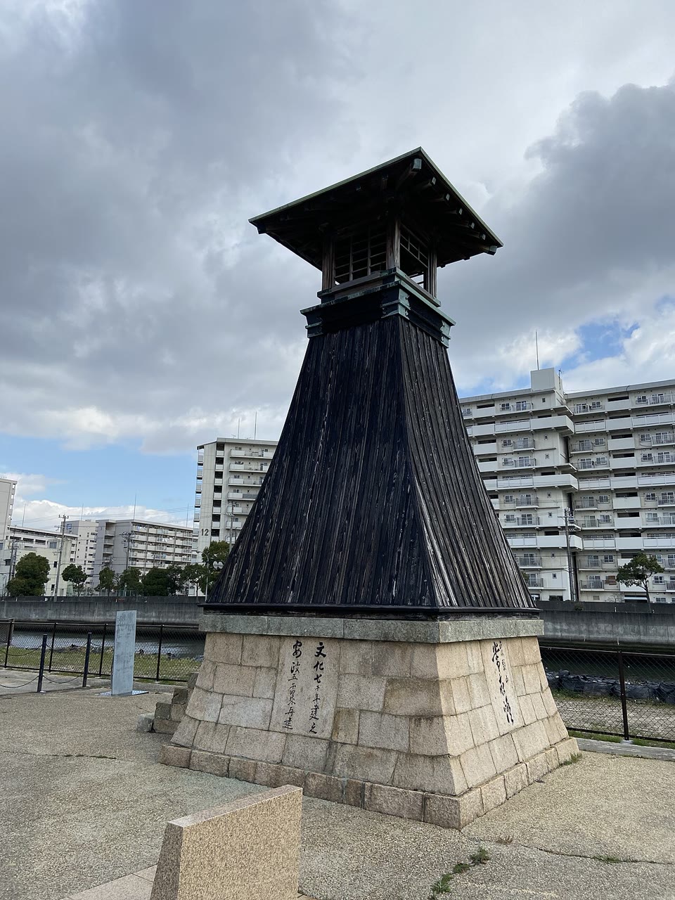 西宮市の今津灯台（2020年撮影）。切石を積んだ台座の上に、黒い板張りで裾の広がった木造の灯台が立ち、頂部に屋根の付いた灯室が載る。台座には「文化七年建之」などの文字が刻まれ、背後に川と集合住宅が見える