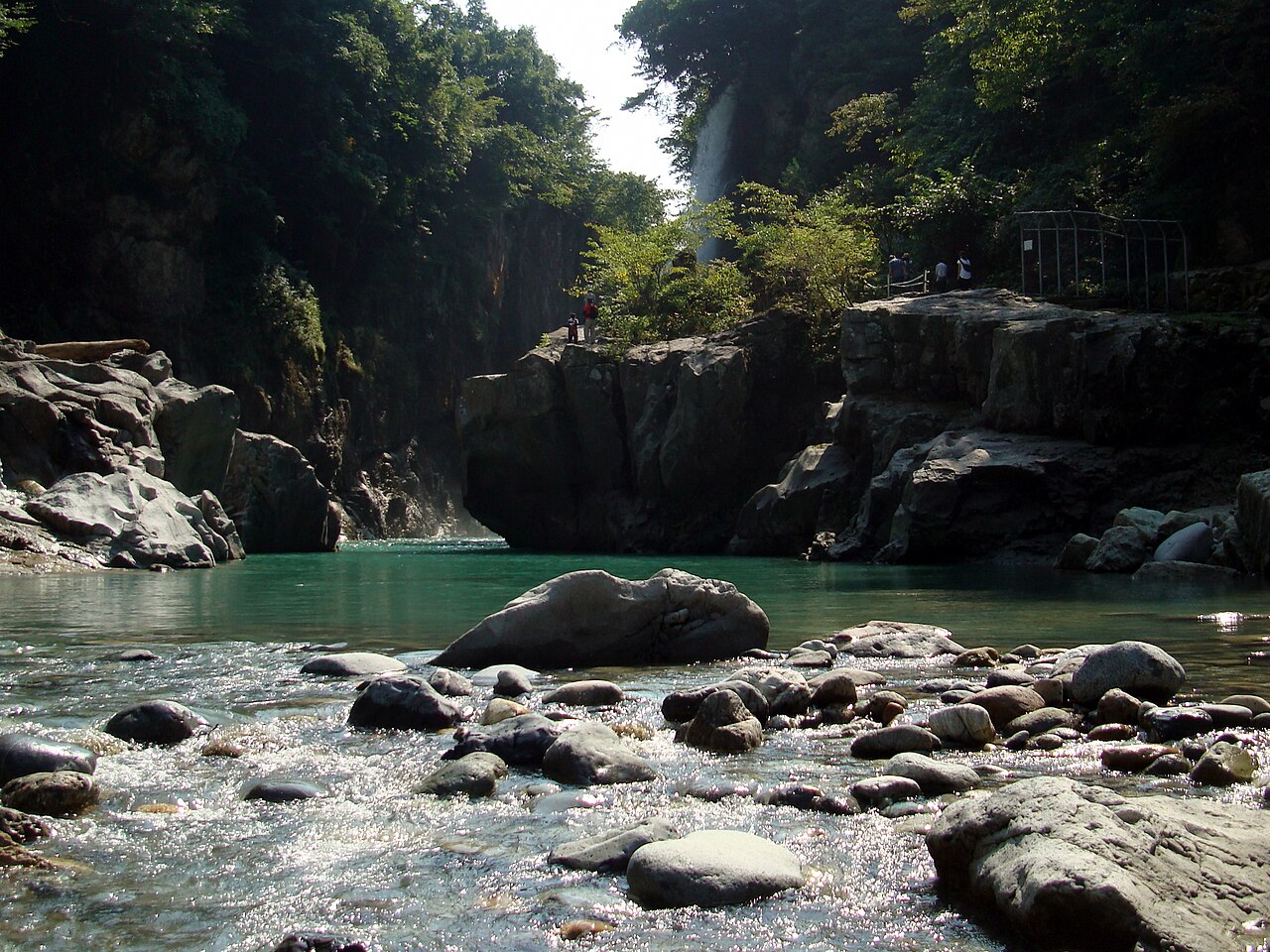 白山市の手取峡谷。緑の木々に覆われた岩の絶壁のあいだを青緑色の手取川が流れ、奥の崖から綿ヶ滝が落ちている。手前の川原には丸い石が並ぶ