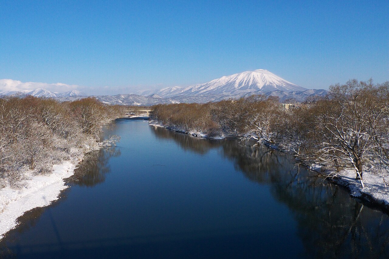 雪の積もった冬の晴れた日の雫石川。青い川面が奥へ延び、両岸には雪をかぶった木々が並ぶ。遠くに雪をいただいた岩手山がそびえる