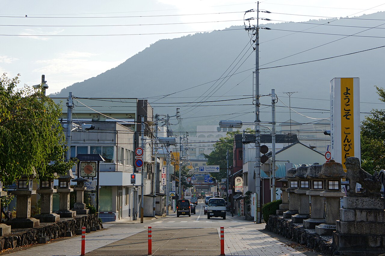 香川県琴平町の駅前の通り。石灯籠が並ぶ道の先に町並みと電線が続き、奥に象頭山がそびえる