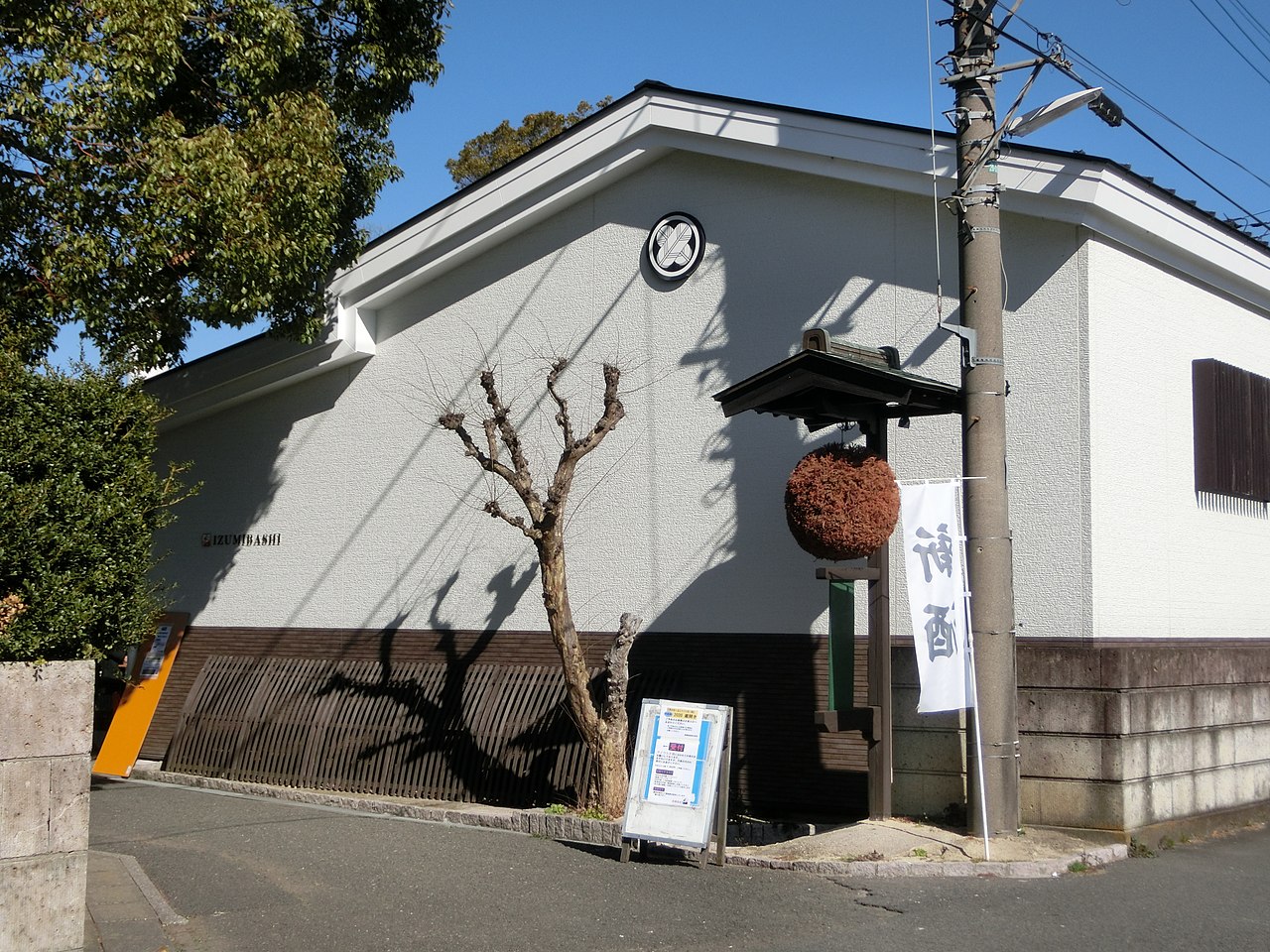 泉橋酒造の本社の建物。白い壁の上部に丸い家紋、壁の左に「IZUMIBASHI」の文字があり、小さな屋根の下に茶色の杉玉が吊るされている。建物の角の前を道路が通り、手前に枝を切り詰めた木が立つ