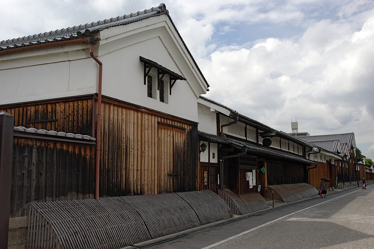 伏見の月桂冠大倉記念館の外観。白壁と焼杉板の蔵が通りに沿って続き、足元には竹を曲げた犬矢来が並ぶ