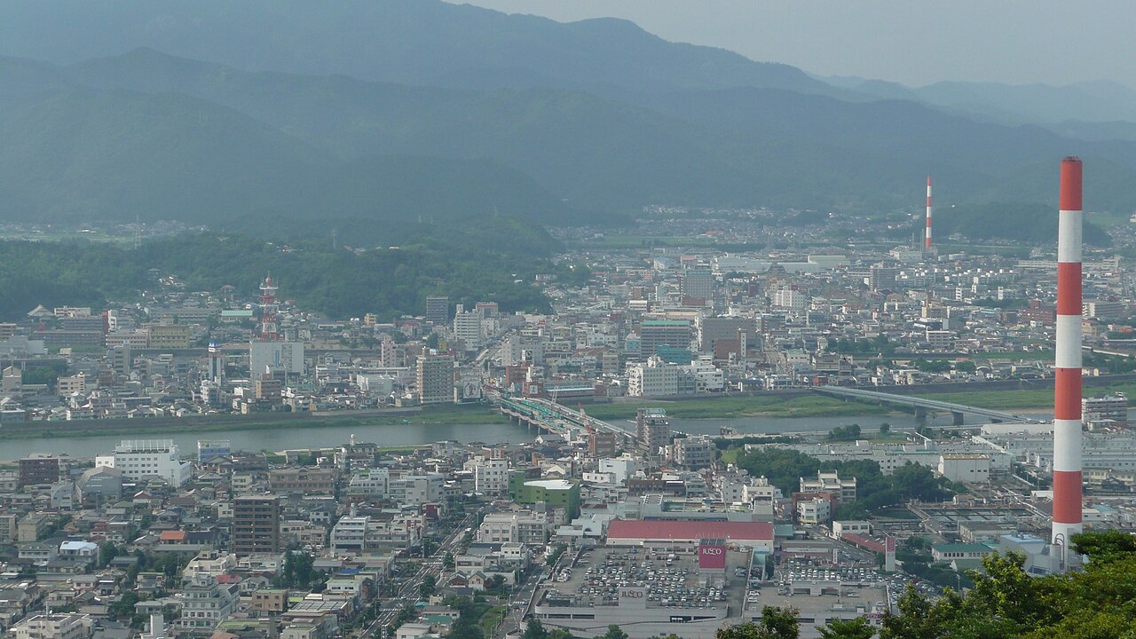 延岡市の愛宕山から見下ろした市街地。手前に建物が並び、中ほどを川が横切り、奥に山並みが連なる。右手前に紅白の煙突が立つ