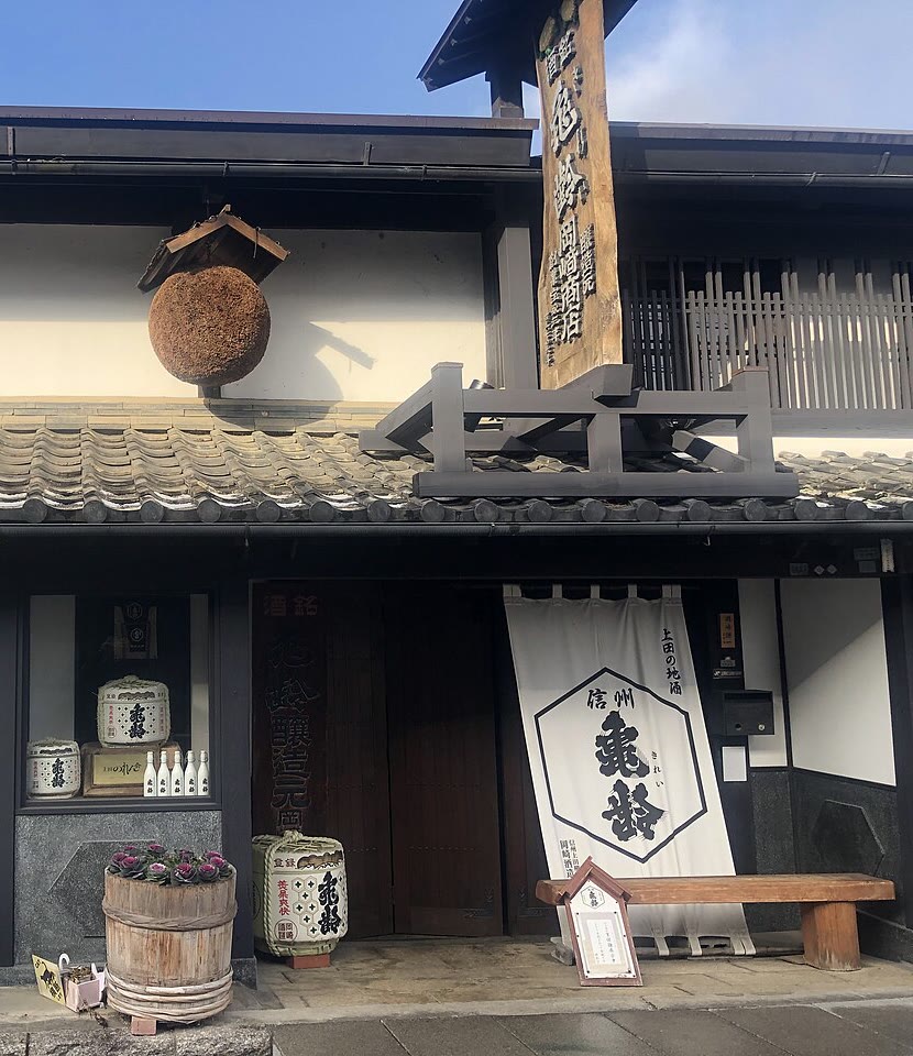 白壁と瓦屋根の岡崎酒造の店先。軒下に杉玉が下がり、入口に「信州亀齢」と書かれた白い暖簾と菰樽が置かれている