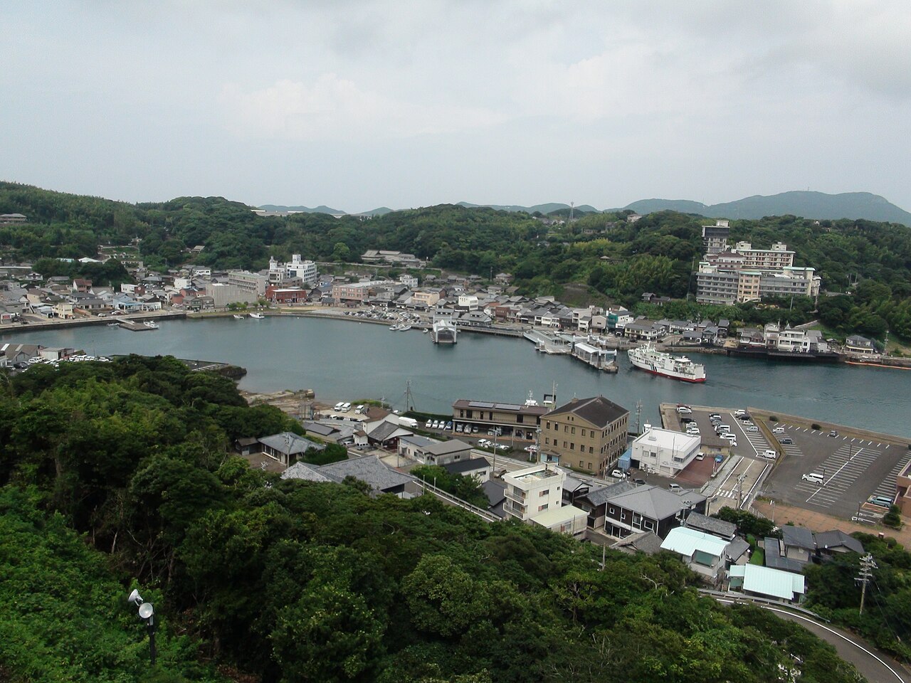 平戸城の高台から見下ろした平戸港。曇り空の下、入り江を囲むように町並みが続き、港には白い船が停まっている。手前は木々の茂る斜面