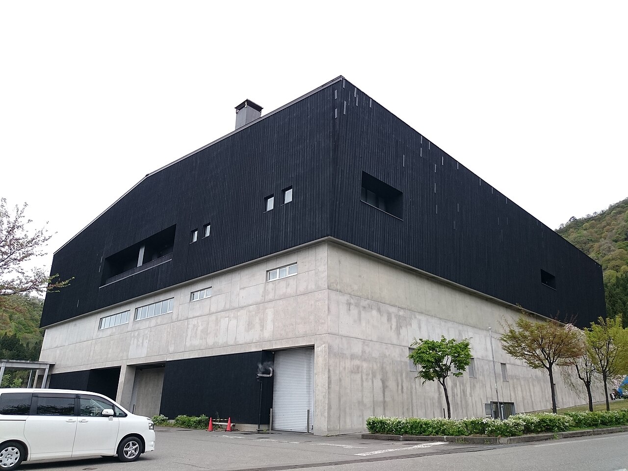 南魚沼市の八海醸造「魚沼の里」にある蔵の外観。コンクリートの基壇の上に黒い板張りの大きな建物が載り、背後に新緑の山が迫る