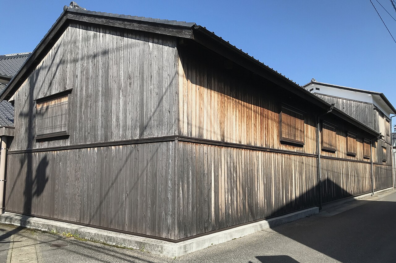鹿島市浜町に建つ富久千代酒造の一号蔵。黒ずんだ板張りの外壁に格子の窓が並び、瓦屋根がかかる。建物の角の前を道路が通っている