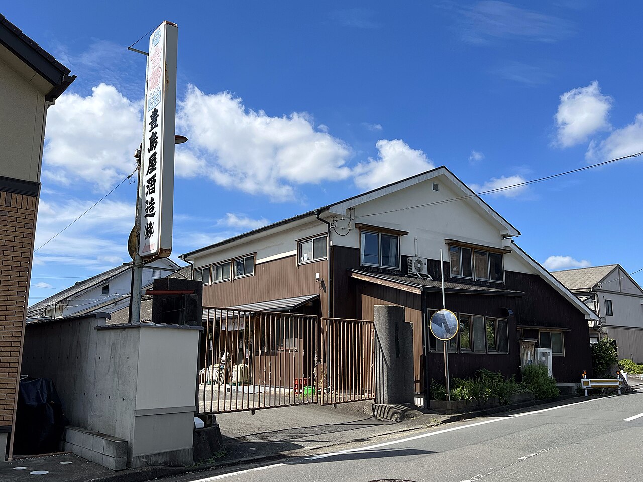 晴れた空の下、道路沿いに建つ豊島屋酒造の門と2階建ての建物。門の脇に「豊島屋酒造(株)」と書かれた縦長の看板が立っている