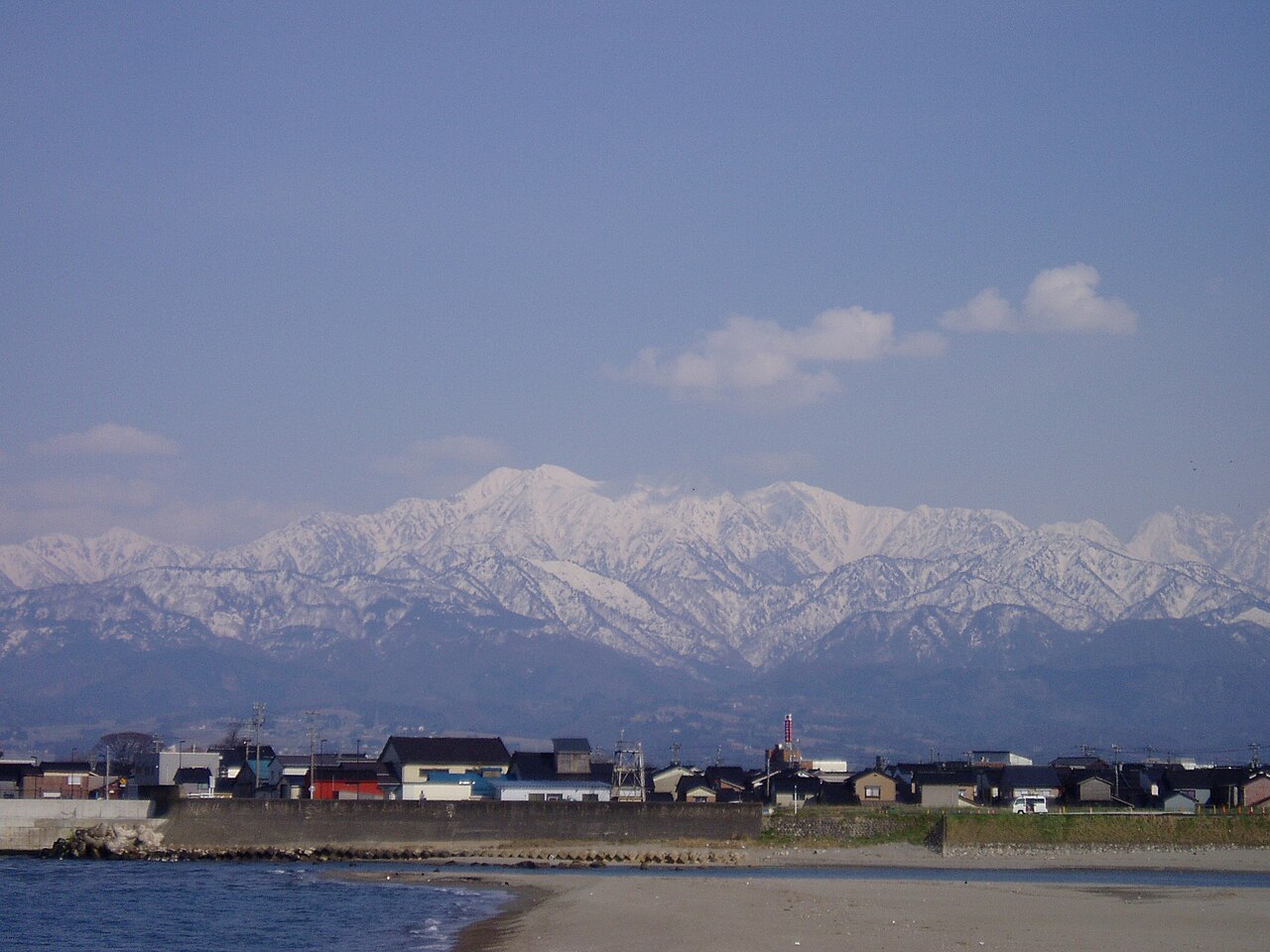 富山市の常願寺川の河口。手前に砂浜と川の水面、その先に低い家並みが続き、奥に雪をかぶった立山連峰の山々が青空の下に連なる