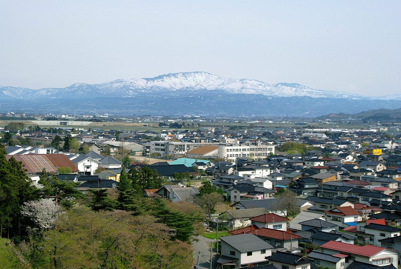 山形県鶴岡市の大山公園から見た、住宅の屋根が広がる鶴岡の市街地と、その奥の平野、雪の残る月山の山並み