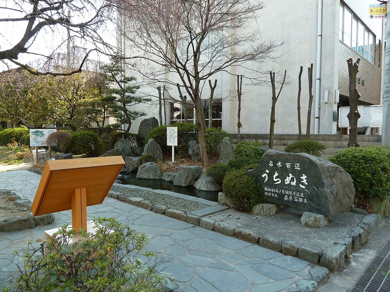 西条市の名水「うちぬき」。「名水百選 うちぬき」と刻まれた石碑の奥に、石で囲まれた水場と植え込みがあり、その後ろに建物が建つ