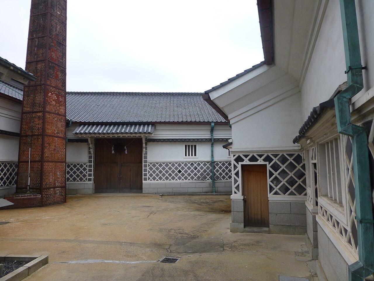 東広島市西条の酒蔵の中庭。左に赤れんがの煙突が立ち、奥と右に、瓦屋根と白壁、下部に白と黒の格子模様の壁をもつ蔵が並ぶ