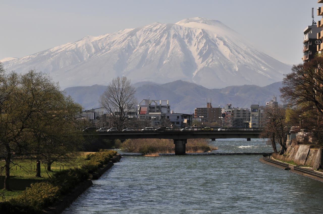 盛岡市の旭橋から見た北上川と岩手山。手前を川が流れ、橋と街並みの向こうに、山頂まで雪をかぶった岩手山がそびえる。岸には芽吹き前の木々