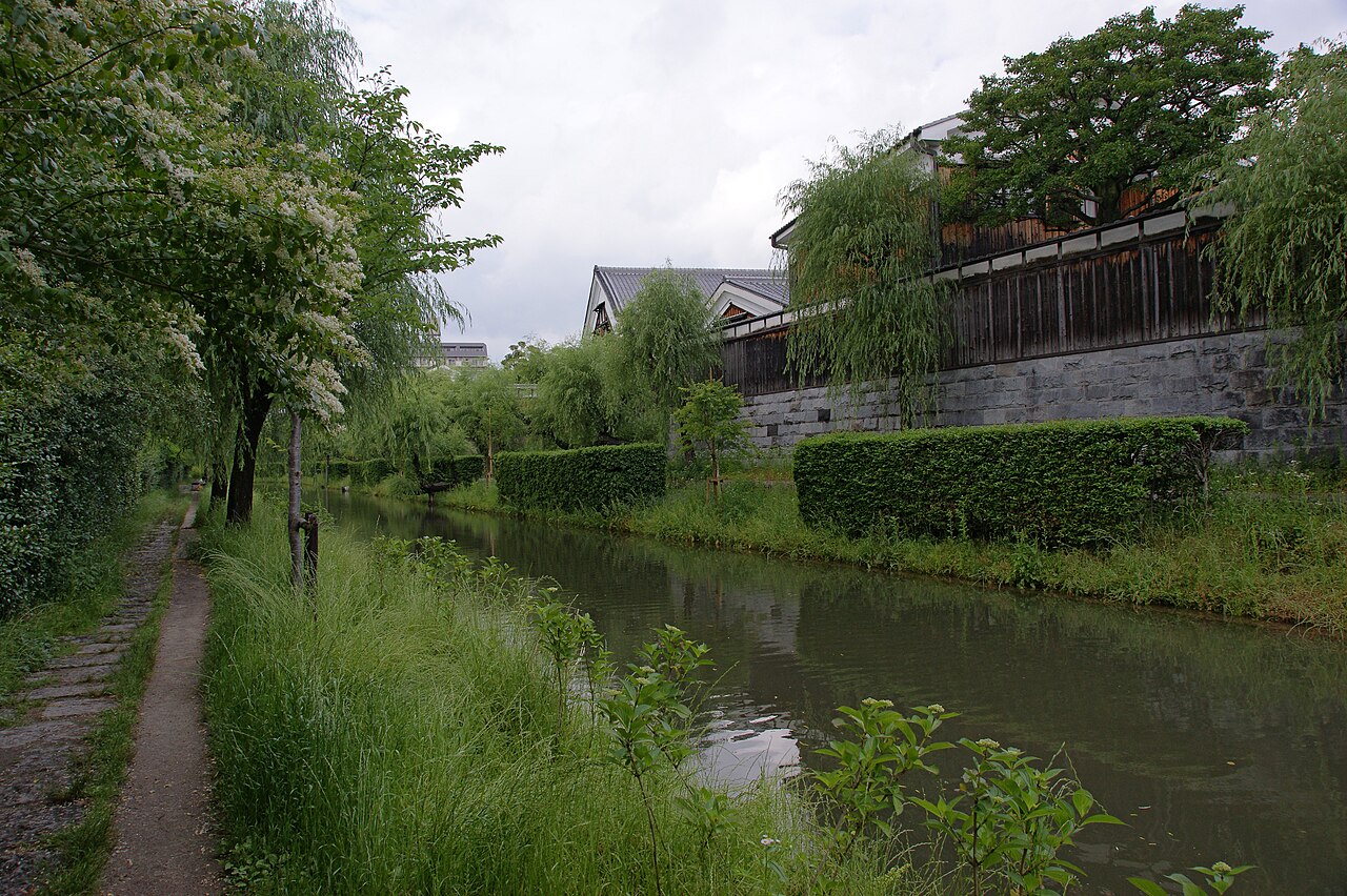 伏見の宇治川派流。柳の並ぶ水路の右岸に、石垣の上に板塀と白壁の酒蔵が続き、左岸には細い遊歩道がある