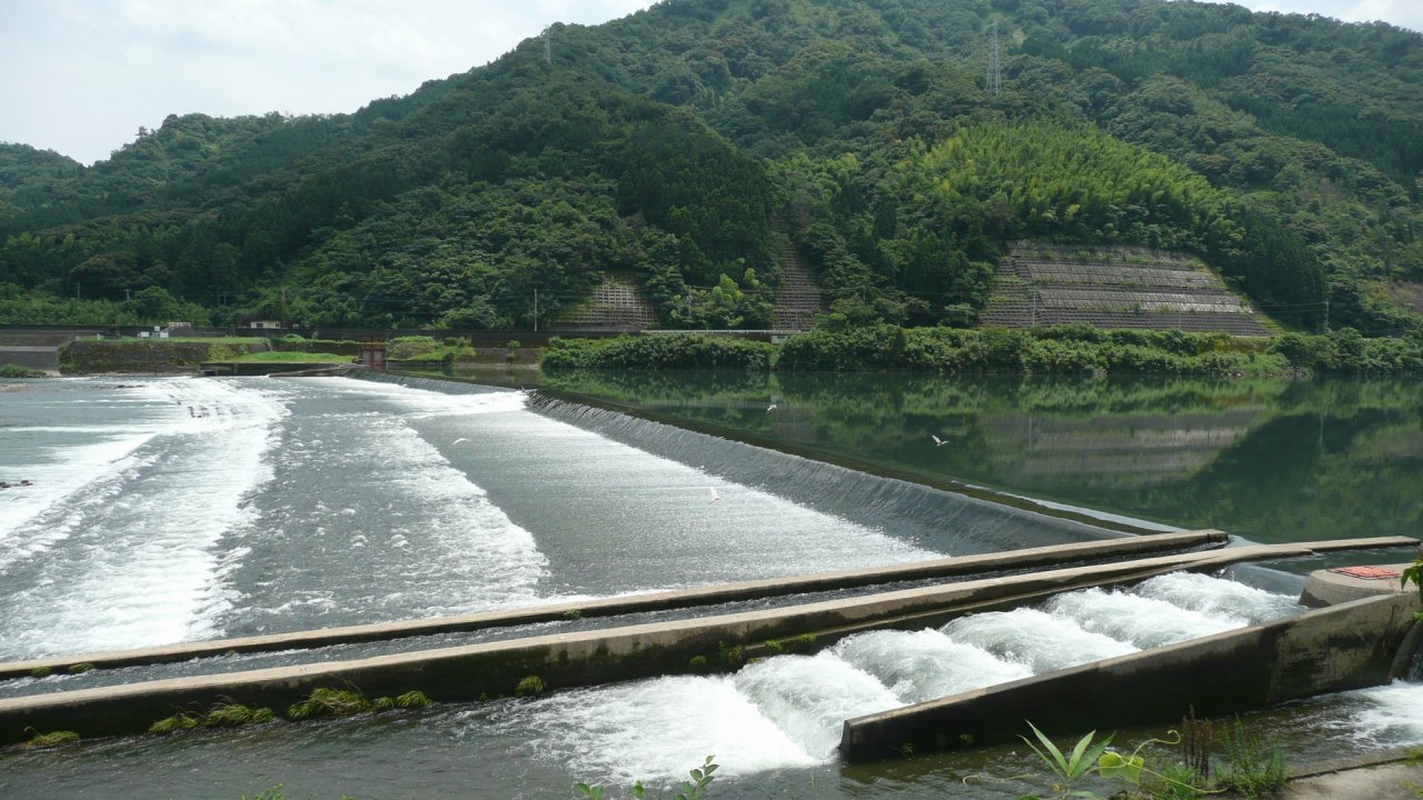 延岡市を流れる五ヶ瀬川の井堰。白く泡立って流れ落ちる水の向こうに、緑の山が川面に映る