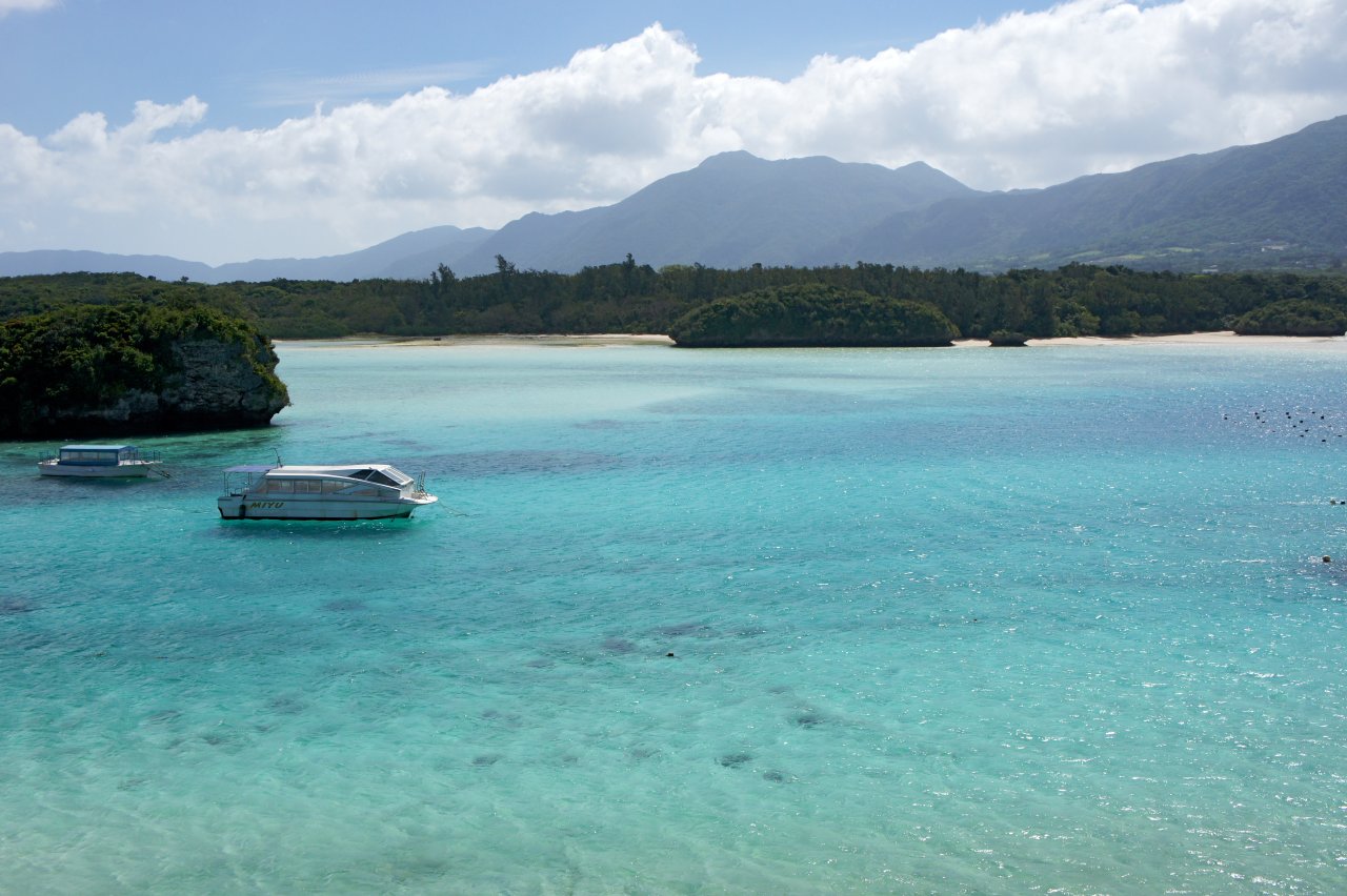 石垣島の川平湾。青く澄んだ浅い海に小さな船が浮かび、緑の森と砂浜の奥に山々が連なる