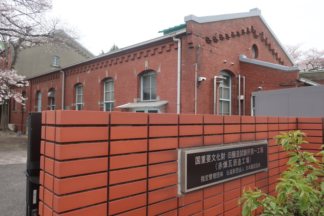北区滝野川の旧醸造試験所第一工場。切妻屋根の赤い煉瓦造りの建物が建ち、手前の煉瓦塀に「国重要文化財 旧醸造試験所第一工場（赤煉瓦酒造工場）」の銘板が付いている。左に桜が咲いている